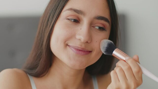 A young Middle Eastern woman smiles while applying makeup at her home. She is engaged in a self-pampering routine, showcasing her beauty skills and enjoying personal care.