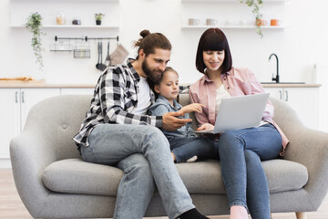 Caucasian family enjoying time together on sofa watching movie on laptop. Young parents with daughter, all smiling, in casual clothing, cozy living room setting.