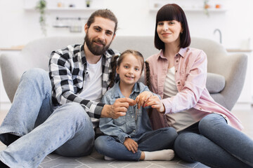 Joyful Caucasian family sitting together holding keys, celebrating new home. Father, mother, and daughter express happiness in cozy living room environment.