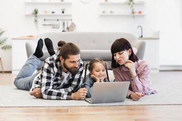 Caucasian family with two adults and child enjoying leisure time together on living room floor using laptop. Child engaged and happy. Comfortable, bright modern setting. Concept of family bonding.