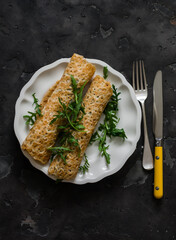 Crepes stuffed with tuna, cucumber, eggs, arugula salad on a dark background, top view