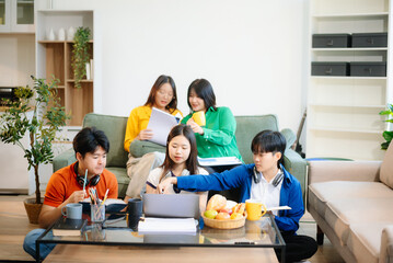 Group of young Asian students working on a project with laptops and tablets. Cozy indoor setting, teamwork,