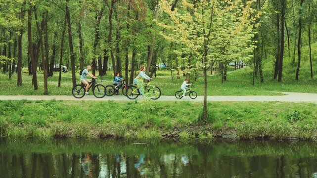 Family of four rides bicycles through green park along pond. Parents and children enjoy outdoor activities and fresh air. Concept: family bonding, healthy lifestyle, nature connection
