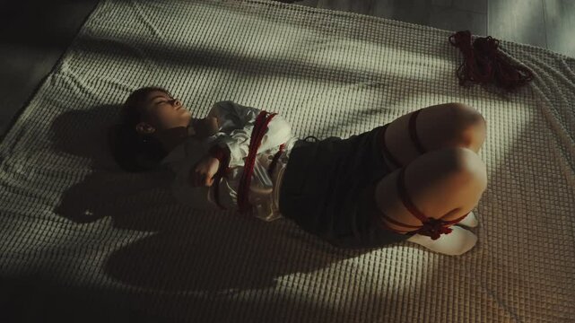 Woman lies on textured rug, bound in intricate red rope shibari. Soft lighting and shadows create intimate, artistic atmosphere