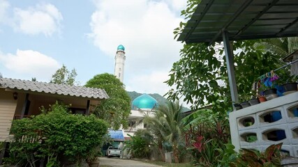 A tranquil setting showcases a mosque with a striking blue dome and tall minaret nestled among lush green trees and houses. The clear sky adds to the serene atmosphere. - Powered by Adobe
