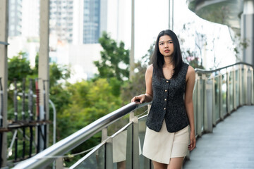 Obraz premium Lifestyle of beautiful Asian female office worker walking on modern pedestrian bridge in urban business district. Wearing stylish black vest and white skirt, looking confident in city setting.
