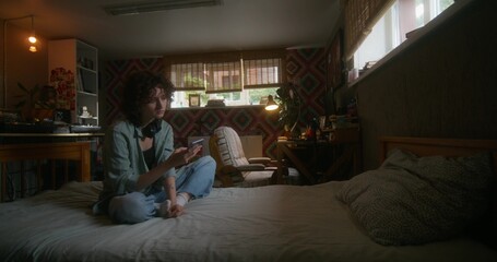 A young woman uses a mobile phone while sitting on a bed at home with a depressed look