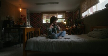 A young woman puts on headphones and uses a mobile phone sitting on the bed. She is upset or...