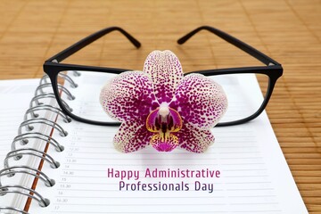 Orchid flower lies on a notebook in front of reading glasses on a wooden background. Selective focus. Greeting card for Secretary day with text 'Happy Administrative Professionals Day.'
