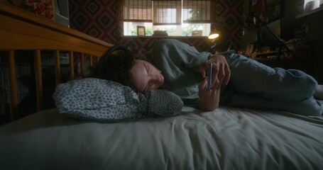 A woman with headphones on her head uses a mobile phone lying in bed at home