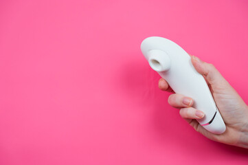 Female hand sticks out white vacuum clitoral vibrator through pink paper background. 