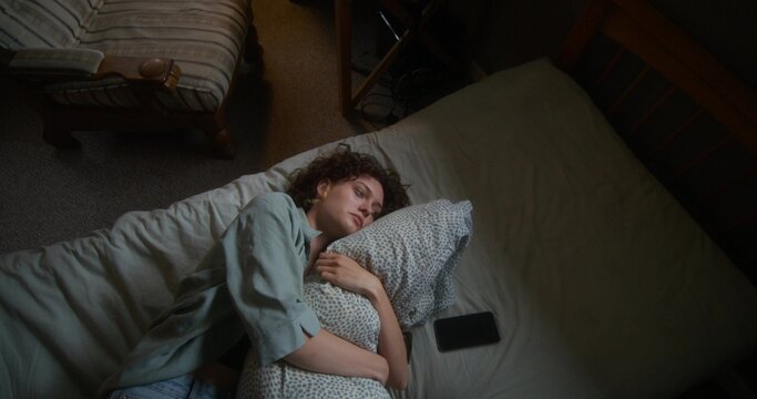 A woman is crying and hugging a pillow lying in bed at home. The mascara is smeared under the eyes. The mobile phone is on her bed