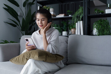 Obraz premium Asian woman sits on sofa listening to music with smartphone in living room