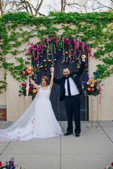 Amidst a beautiful and vibrant floral backdrop, a delighted couple joyfully celebrates their union, marking the special occasion of their wedding with family and friends gathered around them