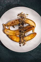 Flambe bananas. Dessert of bananas with pistachios, toasted pumpkin seeds, chocolate and caramel on a white plate on a dark green background. Place for text. Top view. Close-up.