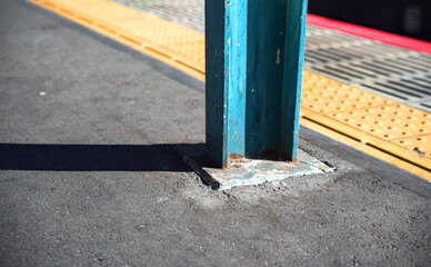 Close-up afternoon view of the base of a weathered blue support beam near the edge of a train station platform, with yellow and red safety lines visible on the side