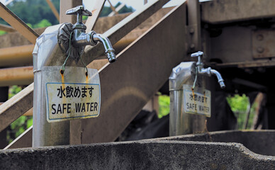 Selective focus on one of two public water faucets next to a metal staircase and platform, on which hang signs that read "safe water" in Japanese and English