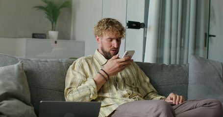 A bearded man in casual clothes records a voice message on a mobile phone sitting on the couch at...