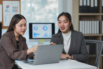 Two professional women engaged in a collaborative work session, discussing data on a laptop in a modern office environment. Charts and graphs visible on a screen in the background