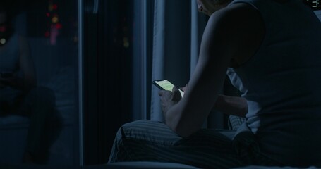 A man uses a mobile phone while sitting on the bed at night. Close-up of his hands, an...