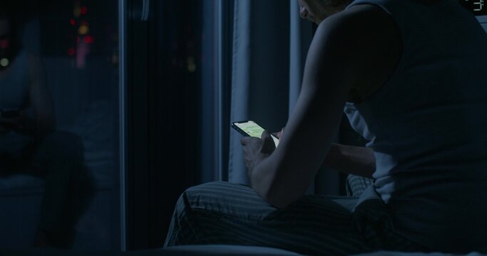 A man uses a mobile phone while sitting on the bed at night. Close-up of his hands, an unrecognizable person. Dark room, home interior