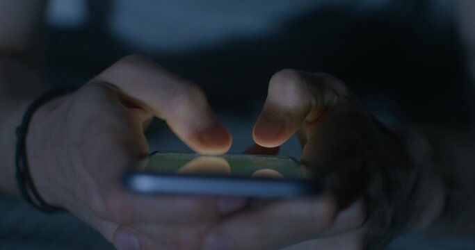 A man uses a mobile phone while sitting on the bed at night. Close-up of his hands, an unrecognizable person. Dark room, home interior
