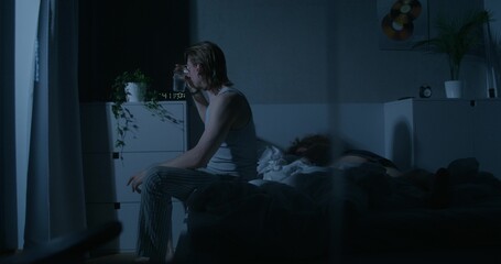 Naklejka premium A man drinks water from a glass and lies down in bed next to his wife. Dark room, home interior