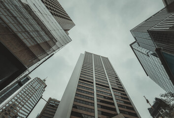 A low-angle, close-up view of towering skyscrapers in Hong Kong, reaching towards an overcast sky. The buildings are a mix of architectural styles, creating a dense urban landscape.