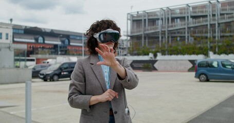 A stylish woman in a jacket wearing a virtual reality headset stands on a city street and moves her...