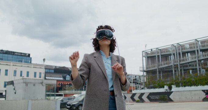 A stylish woman in a jacket wearing a virtual reality headset stands on a city street and moves her arms. Summer day - Powered by Adobe