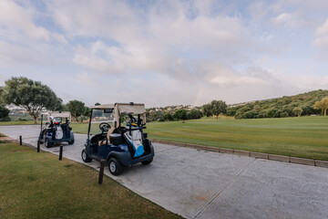 Two golf carts on a paved path by a lush golf course with trees, a distant rainbow, and rolling...