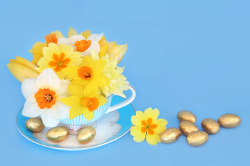Easter flowers in tea cup with gold chocolate eggs. Abstract surreal holiday seasonal composition on blue background.