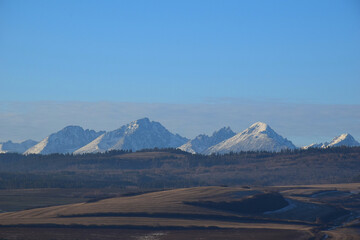 Tatras mountains in Slovakia rise majestically, their snow-capped peaks glistening under a crisp blue sky, creating a serene yet dramatic contrast with the rolling hills and dense forests below. 