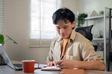 A smart Asian businessman is contemplating his work ideas and writing them in a book.