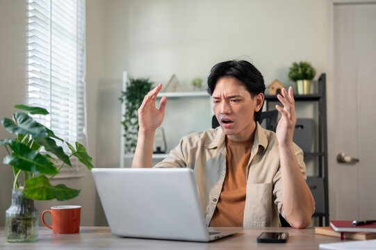 Angry Asian businessman sits at his desk, looking at his laptop screen with a frustrated expression.