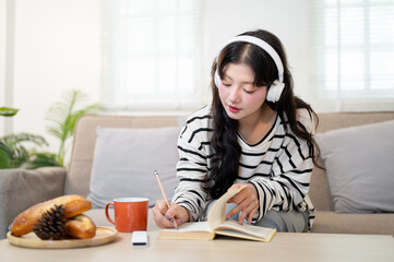 A beautiful, relaxed Asian woman is writing in her diary while listening to music in the living room