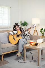 Calm Asian woman is deep in thought as she listens to music through her headphones and plays guitar.