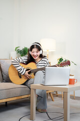 A cheerful Asian woman is live on her social media while playing guitar on a sofa at home.