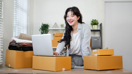 A smiling, happy Asian female online business entrepreneur is at her desk, working on her laptop.