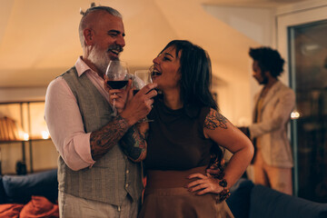Mature couple with tattoos enjoying wine at a social gathering - Stylish tattooed couple sharing a joyful moment with wine glasses at an evening party indoors