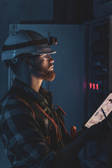 Electrician in overalls, focused on work in switchboard with fuses, using tablet.