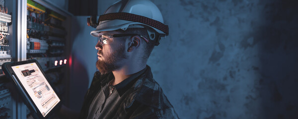 A male electrician in overalls, focused on work in switchboard with fuses, using tablet.