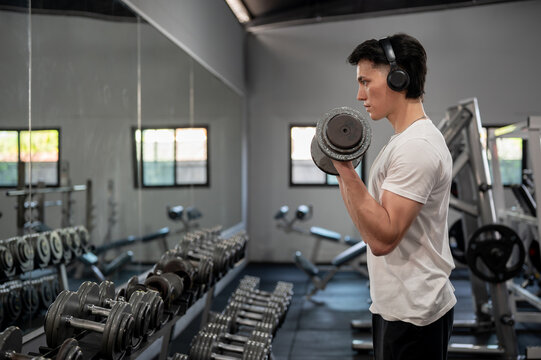 Focused Asian man is working out with a dumbbell in front of the dumbbell rack and mirror in the gym