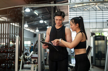 An Asian male fitness instructor is adjusting a workout program with a female client in the gym.
