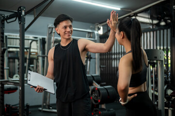A cheerful, athletic Asian male fitness trainer is giving a high five to his client in the gym.