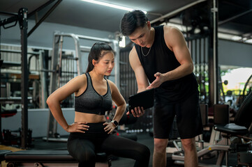 A female client sits on a fitness bench, discussing her workout program with her fitness trainer.
