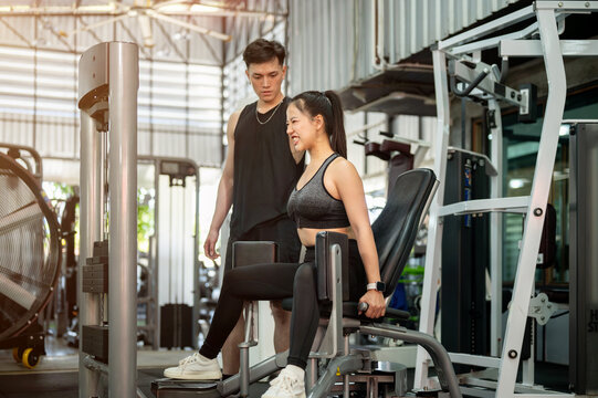 A determined Asian woman is being trained by a fitness coach, using the adductor machine in the gym.