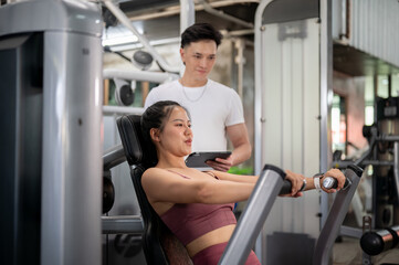 A fit Asian woman is being instructed by a professional fitness trainer, using a chest press machine