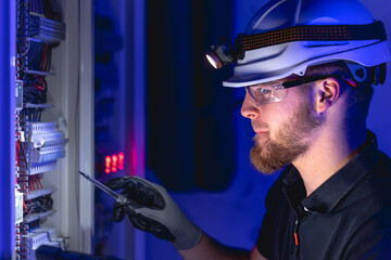 A male electrician works in a switchboard in overalls against the backdrop of emergency lighting.