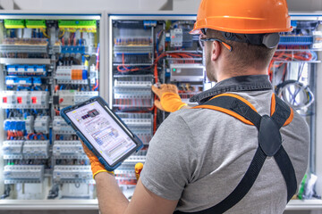 A male electrician in overalls, focused on work in switchboard with fuses, using tablet.